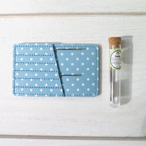 May include: A light blue needle holder with white polka dots and two needles. A clear glass vial with a cork stopper and a label that reads "Tulip Embroidery Needles Assorted" is next to the holder. The background is a white wooden surface.