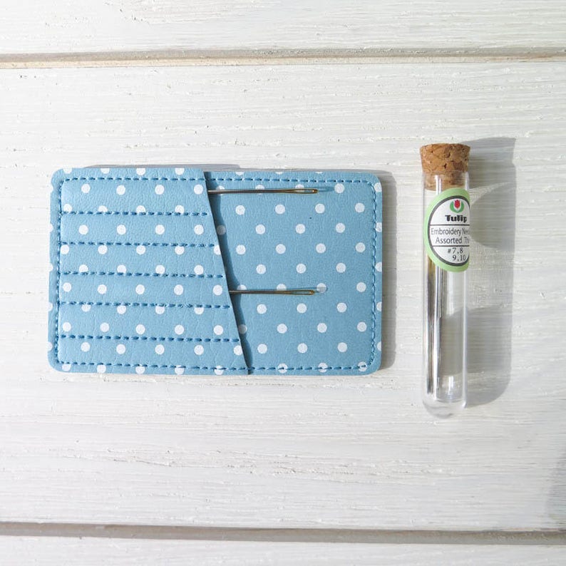 May include: A light blue needle holder with white polka dots and two needles. A clear glass vial with a cork stopper and a label that reads "Tulip Embroidery Needles Assorted" is next to the holder. The background is a white wooden surface.