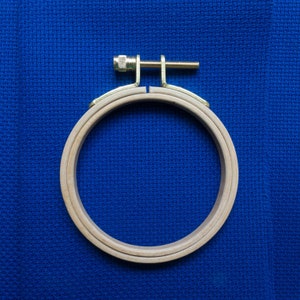 May include: A circular wooden embroidery hoop with a brass screw and bracket, set against a bright blue fabric background. The hoop is made of light-coloured wood and is designed to hold fabric taut for cross-stitch or embroidery projects.