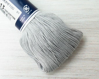 Puede incluir: Un ovillo de hilo de algodón gris, con texto japonés y el número 13 en la etiqueta. La etiqueta también indica "100% Algodón" y "Made in Japan". El hilo está bien enrollado, adecuado para diversos proyectos de costura.