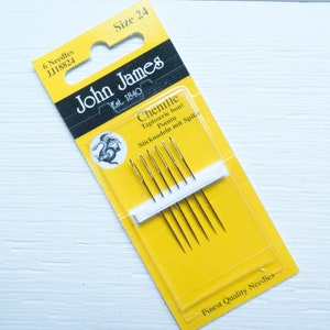 May include: A package of John James chenille needles, size 24, with the text "6 Needles" and "Finest Quality Needles". The yellow and black packaging displays the brand logo and product details. The package contains six needles.