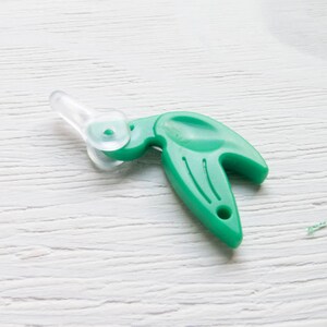 May include: A green plastic yarn cutter tool with a clear plastic blade and handle. The tool has a curved handle with a hole at the end and a small blade for cutting yarn or thread. The tool is designed for crafting and sewing projects.