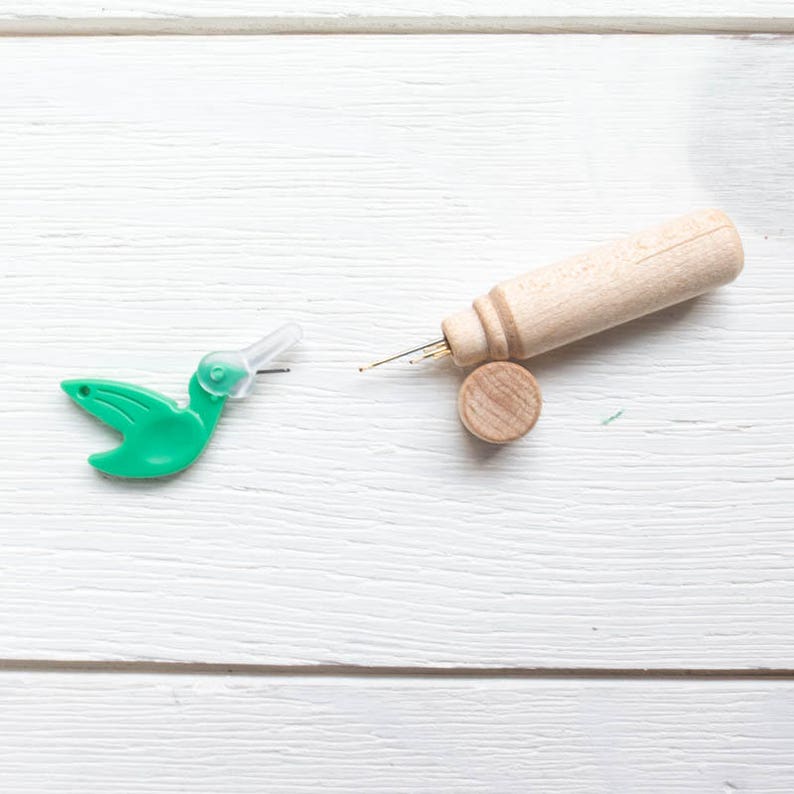 May include: A green plastic hummingbird needle threader and a wooden punch needle tool with a small wooden disc. The needle tool has a light brown wooden handle and a metal needle. The hummingbird threader is bright green.