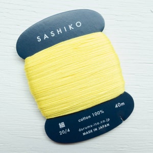 May include: A spool of bright yellow Sashiko cotton thread. The thread is wrapped around a dark blue label with the word "SASHIKO" in white. The label also includes text indicating it is 100% cotton, 40m, and made in Japan.
