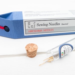 May include: A blue box of assorted sewing needles with the text "Sewing Needles Assorted" and "Hiroshima Needles". A cork stopper and a clear tube of needles are also present.
