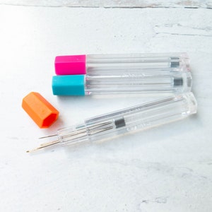 May include: Clear plastic needle cases with colorful caps in pink, teal, and orange. The cases contain multiple needles, ideal for sewing or crafting projects. The needles are visible inside the transparent tubes.