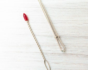 May include: Two silver skin care tools on a white wooden surface. One tool has a red tip and a twisted handle. The other tool is straight with a loop and a small opening on the end. Both tools are designed for skincare.