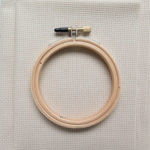 May include: A wooden embroidery hoop with a brass screw and black cap, set against a neutral-toned fabric background. The hoop is made of two concentric rings, designed to hold fabric taut for cross-stitch or embroidery projects.