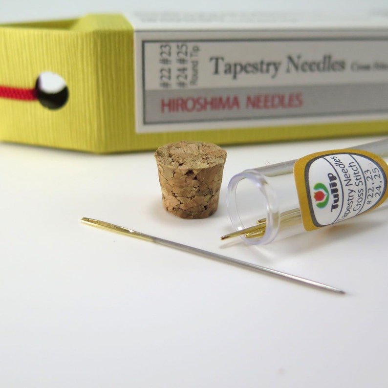 May include: A close-up of tapestry needles and accessories. A single needle lies in front of a cork stopper and a clear plastic tube containing several gold-tipped needles. The packaging reads "Tapestry Needles" and "Hiroshima Needles."