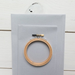 May include: A light gray cross-stitch fabric with a wooden embroidery hoop. The hoop is secured with a brass screw and black hardware. The fabric is layered with two pieces of different sizes.