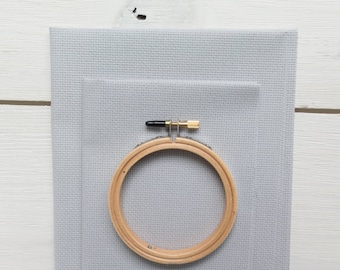 May include: A light gray cross-stitch fabric with a wooden embroidery hoop. The hoop is secured with a brass screw and black hardware. The fabric is layered with two pieces of different sizes.