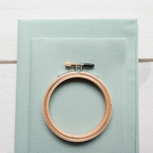 May include: A wooden embroidery hoop with a brass screw and black tip rests on two pieces of light green cross-stitch fabric. The fabric is stacked on top of each other, creating a layered effect. The background is a white wooden surface.