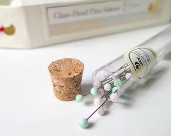 May include: A collection of glass-head pins with light pink and mint green heads, spilling from a clear tube. A cork stopper sits beside the tube. The packaging box in the background reads "Glass-Head Pins Sakura".