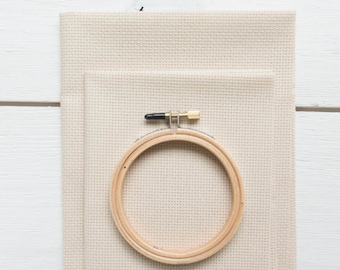 May include: A cross-stitch kit featuring a wooden embroidery hoop, beige Aida cloth, and a white hanging loop. The hoop is secured with a black and gold metal screw. The cloth is folded and layered.