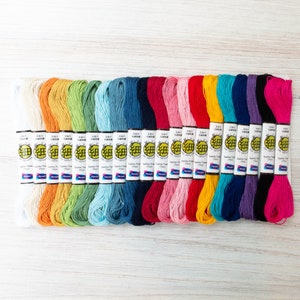 May include: A collection of colorful embroidery floss in a variety of shades, including white, orange, green, blue, pink, red, yellow, and purple. Each spool is labeled with Japanese text and the brand name "Olympus". The floss is neatly arranged on a light wood surface.