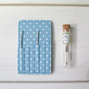 May include: A light blue needle case with white polka dots and several pockets, holding two needles. A clear glass vial with a cork stopper contains embroidery needles, with the text "Tulip" and "Embroidery Needle Assorted Thin".