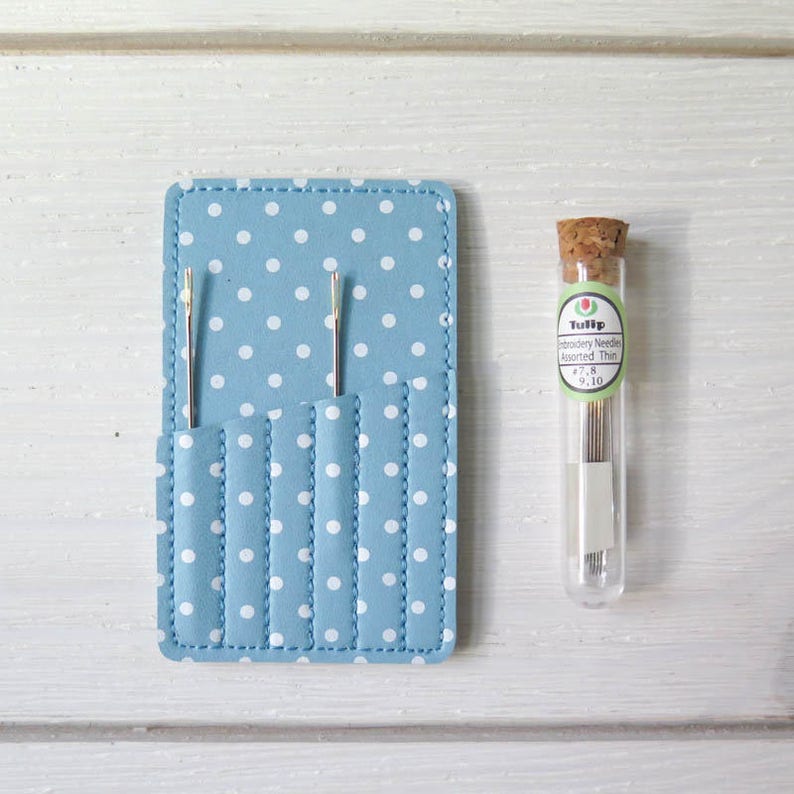 May include: A light blue needle case with white polka dots and several pockets, holding two needles. A clear glass vial with a cork stopper contains embroidery needles, with the text "Tulip" and "Embroidery Needle Assorted Thin".