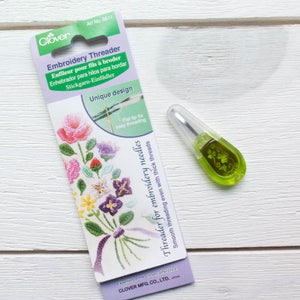 May include: An embroidery threader tool with a green handle and a clear plastic cover, next to a package with floral embroidery designs. The package reads "Embroidery Threader" and "Unique design".