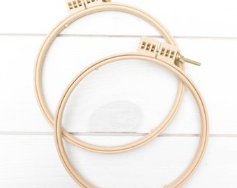 May include: Two beige embroidery hoops, one slightly overlapping the other, against a white wooden background. Each hoop has a tightening mechanism with a screw. The hoops are circular and made of a light-colored wood or plastic.