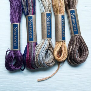 May include: Five skeins of Cosmo Seasons embroidery floss in shades of purple, blue, and brown. Each skein is labelled with "Seasons" and "100% Cotton Made in Japan". The floss is displayed on a light blue surface.