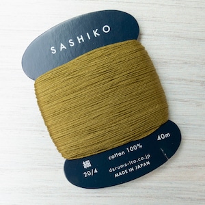 May include: A spool of olive green Sashiko cotton thread. The dark blue label reads "SASHIKO" in white. The spool contains 40m of 100% cotton thread, ideal for embroidery and sewing. Made in Japan.