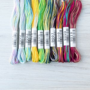 May include: A collection of colorful embroidery floss skeins, each with a white label bearing a number. The floss colors range from pastel blues and greens to vibrant yellows, greens, pinks, and reds. The skeins are arranged on a light-colored wooden surface.