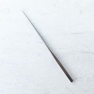 May include: A long, slender, stainless steel tool with a pointed tip. The tool has a flat, rectangular handle and tapers to a fine point. The object is set against a white background.
