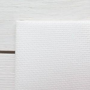 May include: Close-up of a white, textured fabric, possibly a towel or cloth, with a woven pattern. The fabric is folded over, and the background is a white wooden surface.