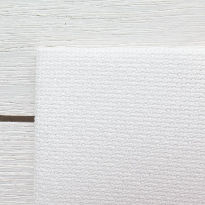 May include: Close-up of a white, textured fabric, possibly a towel or cloth, with a woven pattern. The fabric is folded over, and the background is a white wooden surface.
