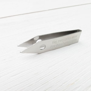 Puede incluir: Pinzas de acero inoxidable con punta fina. El texto "THE TWEEZERS WITH PIN-POINT-PRECISION" est&aacute; grabado en el lateral. Las pinzas se encuentran sobre un fondo de madera blanca.