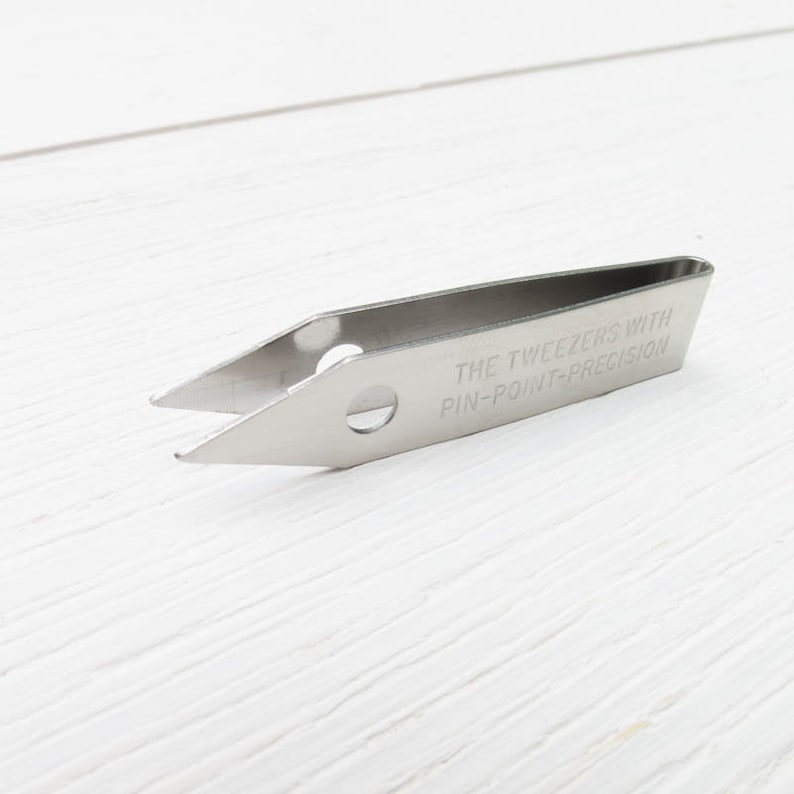 Puede incluir: Pinzas de acero inoxidable con punta fina. El texto "THE TWEEZERS WITH PIN-POINT-PRECISION" est&aacute; grabado en el lateral. Las pinzas se encuentran sobre un fondo de madera blanca.