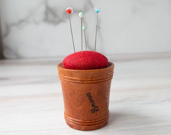 Puede incluir: Un cojín de alfileres de madera con una parte superior de tela roja, que contiene varios alfileres con cabezas de vidrio de colores. La base de madera es cilíndrica con un diseño decorativo y la palabra "nore" es visible en el lateral.