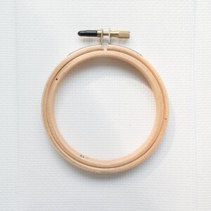 May include: A circular wooden embroidery hoop with a brass screw and black plastic tightening mechanism. The hoop is made of light-coloured wood and is designed to hold fabric taut for needlework projects.