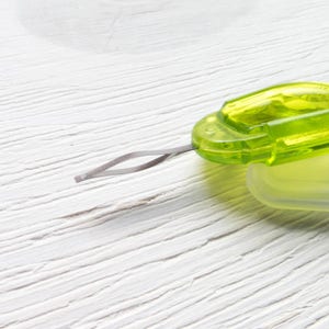 May include: A close-up of a green and clear plastic sewing tool with a silver metal needle. The tool has a rectangular shape with rounded edges and a small loop at the end of the needle. The background is a white wooden surface.