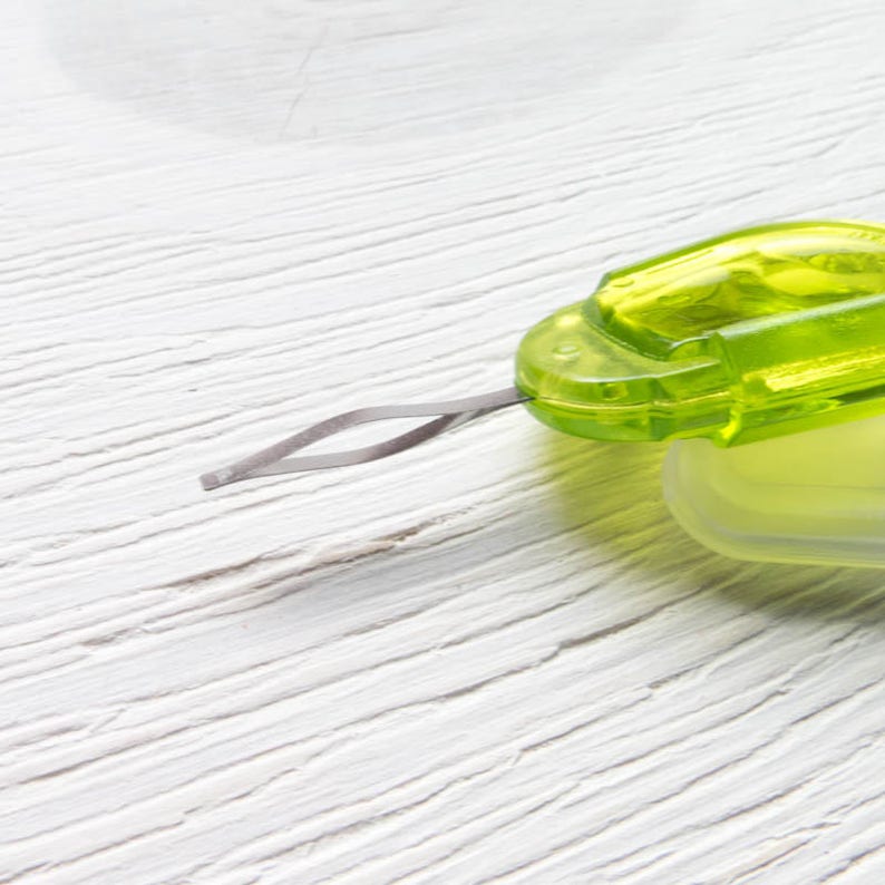 May include: A close-up of a green and clear plastic sewing tool with a silver metal needle. The tool has a rectangular shape with rounded edges and a small loop at the end of the needle. The background is a white wooden surface.