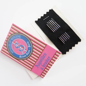 May include: A package of embroidery needles and a decorative box. The box has pink and white stripes with text and a blue and pink emblem. The needles are silver and are on a black fabric.