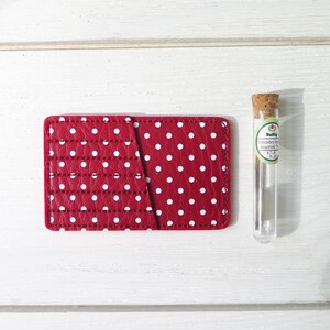 May include: A red, polka-dotted card holder with white stitching and a diagonal pocket. Next to it is a glass vial with a cork stopper, labeled "Tulip Embroidery Needles Assorted Sizes".