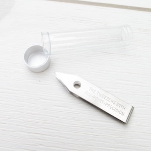Puede incluir: Una pinza de acero inoxidable con punta y un peque&ntilde;o orificio circular. El texto "THE TWEEZERS WITH PIN-POINT-PRECISION" est&aacute; grabado en el lateral. Tambi&eacute;n hay un tubo de pl&aacute;stico transparente y una tapa blanca.