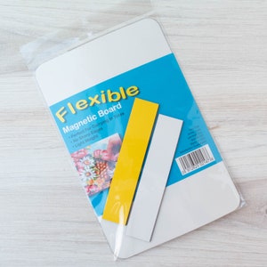 May include: A white flexible magnetic board with a blue and yellow label, packaged in clear plastic. The label reads "Flexible Magnetic Board". Two rectangular magnets, one yellow and one white, are included.