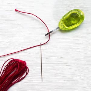 May include: A close-up of sewing tools on a white surface. A bundle of red thread, a needle, and a green thread cutter are visible. The needle is positioned vertically, and the thread is cut by the thread cutter.