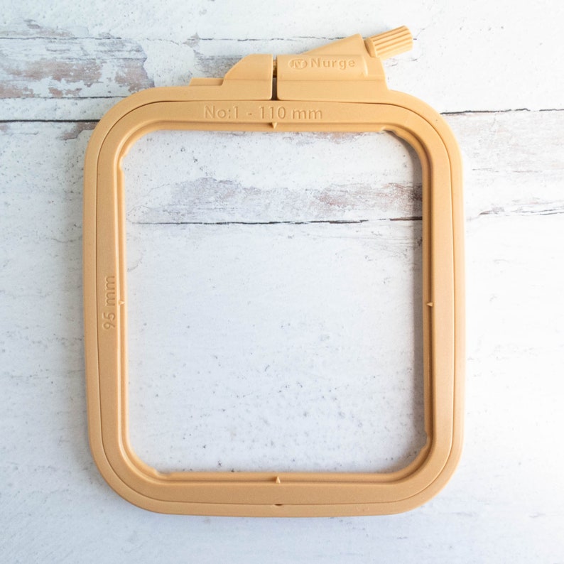 May include: A tan-colored embroidery hoop with the text "No:1 - 110 mm" and "95 mm" printed on it. The hoop is made of plastic and has a rectangular shape with rounded corners. The brand name "Nurge" is also visible.