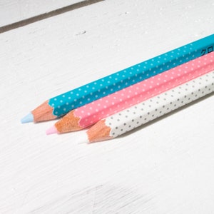 May include: Three colored pencils with polka dot patterns in turquoise, pink, and white. The pencils have light-colored tips and are arranged diagonally on a white wooden surface. The pencils are sharpened and ready for use.