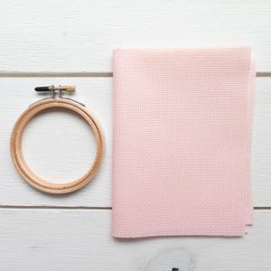 May include: A wooden embroidery hoop with a metal clasp next to a folded piece of light pink cross-stitch fabric. The fabric has a grid-like texture, and the hoop is round.