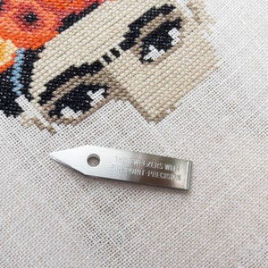 May include: A close-up of a cross-stitch project with a butterfly design in shades of orange, beige, and black. A silver tweezer with the text "THE TWEEZERS WITH PIN-POINT-PRECISION" is in the foreground, suggesting a craft or hobby theme.