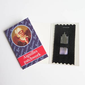 May include: A sewing needle set with a booklet featuring a vintage illustration of a woman in a traditional bonnet. The booklet has the text "Aiguilles patchwork" on a red banner. Needles are displayed on a black fabric.