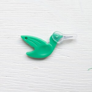 May include: A green plastic hummingbird-shaped tool with a clear, pointed tip. The tool has a small hole near the wing area and is designed with a curved body and a long, slender beak. The object is set against a white, textured background.