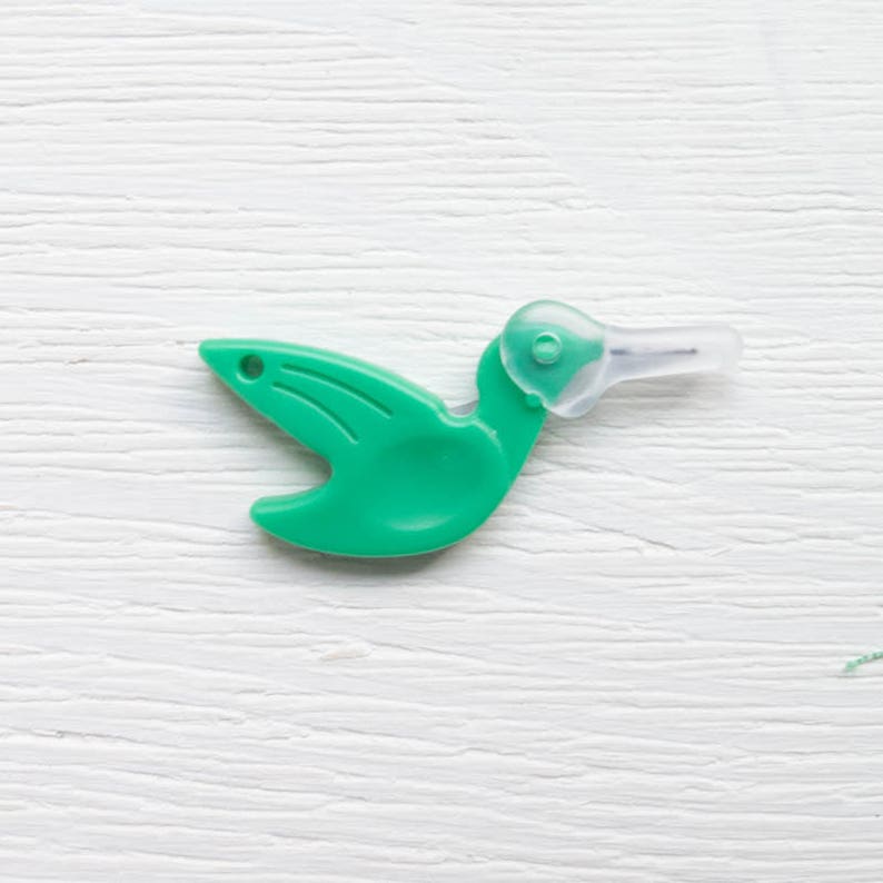May include: A green plastic hummingbird-shaped tool with a clear, pointed tip. The tool has a small hole near the wing area and is designed with a curved body and a long, slender beak. The object is set against a white, textured background.
