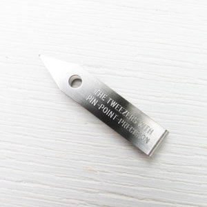Puede incluir: Una pinza de acero inoxidable con punta y un agujero circular. El texto "THE TWEEZERS WITH PIN-POINT-PRECISION" est&aacute; grabado en el cuerpo rectangular de la herramienta. La pinza est&aacute; dise&ntilde;ada para trabajos de precisi&oacute;n.