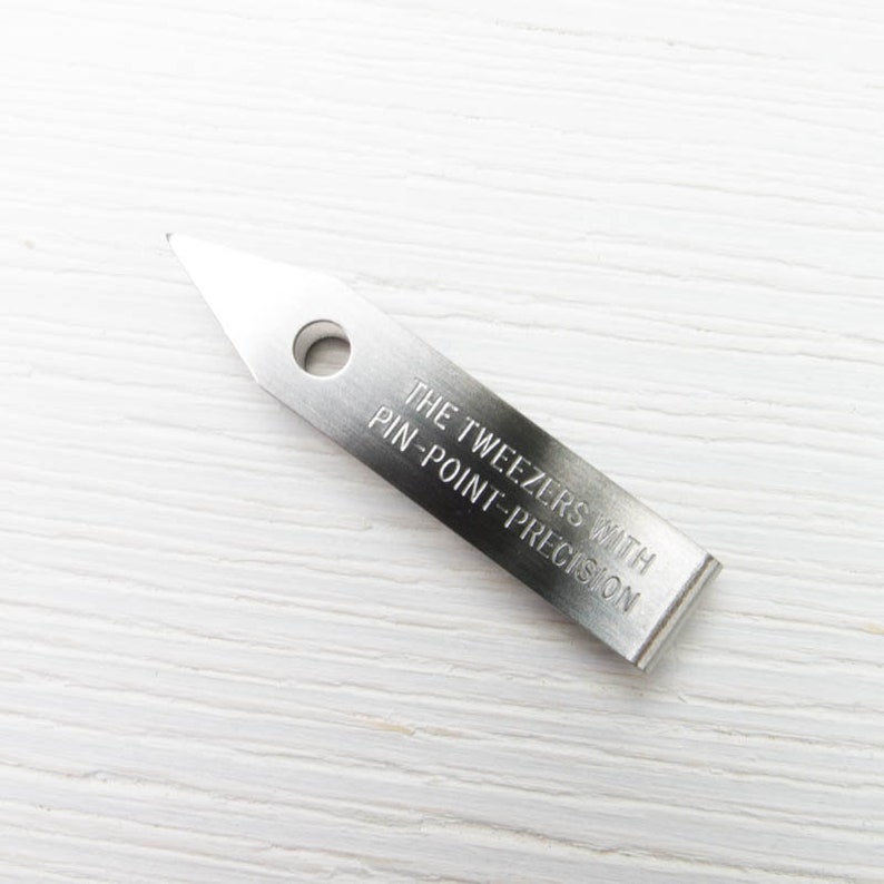 Puede incluir: Una pinza de acero inoxidable con punta y un agujero circular. El texto "THE TWEEZERS WITH PIN-POINT-PRECISION" est&aacute; grabado en el cuerpo rectangular de la herramienta. La pinza est&aacute; dise&ntilde;ada para trabajos de precisi&oacute;n.