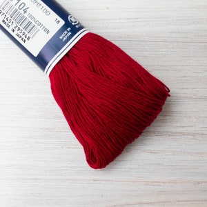 May include: A skein of vibrant red 100% cotton embroidery floss. The label reads "104" and "Made in Japan". The floss is tightly wound, showcasing its rich color and texture. Ideal for various needlecraft projects.